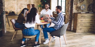 Modern kreatif stüdyoda üniversite projesi üzerinde çalışırken gündelik kıyafetlerle masada dizüstü bilgisayar ve kağıtlarla tam vücut neşeli öğrenciler.