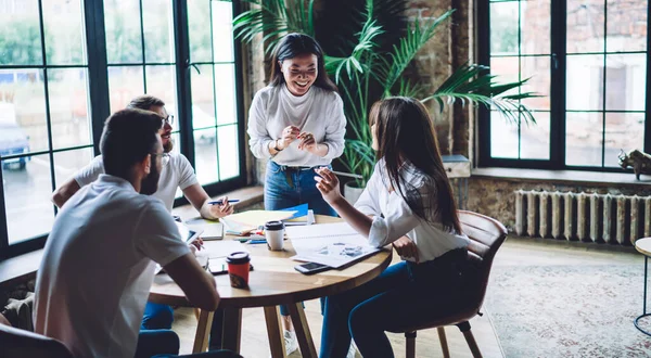 Happy young multiethnic team members wearing jeans and white shirts gathering at round table with papers and discussing new ideas while working together in contemporary light workplace