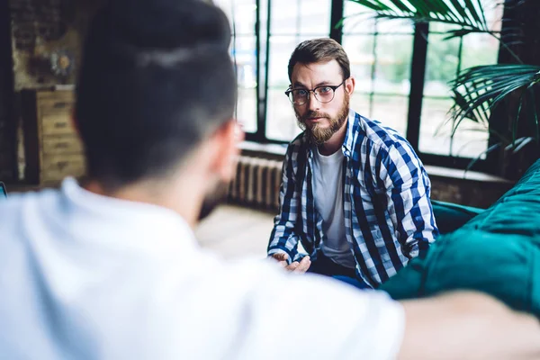 Modern gözlüklü ciddi sakallı bir adam iş arkadaşıyla konuşuyor. Gün ışığında rahat bir iş yerinde proje üzerinde birlikte çalışıyorlar.