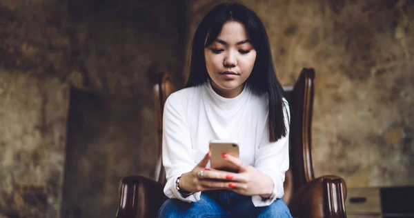 在简约的房间里 一个人坐在皮革扶手椅上 穿着休闲装的亚洲黑发女子的低角度浏览智能手机 — 图库照片
