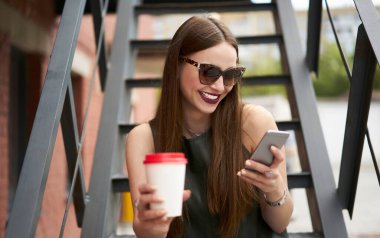 Şık güneş gözlüklü neşeli kadın milenyumu cep telefonuyla internet metinlerini tarıyor ve internetten rezervasyon yaptırıyor, kafkasyalı mutlu turist kahvesiyle ağlarda komik yayınlar okuyor.