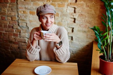 Yüksek açılı, şık giyinen, dişlek gülüşlü, ahşap masaya oturup kafeteryada kahve içen iyimser bir kadın.