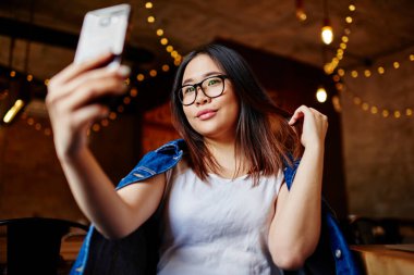 Kafeteryada oturmuş cep telefonuyla selfie çekerken saçlarına dokunan sıradan giysili Asyalı bir kadın.