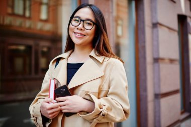Genç, dişlek gülüşlü ve uzun saçlı bir kadın. Şehir caddesinde cep telefonu ve not defteriyle kameraya bakıyor.