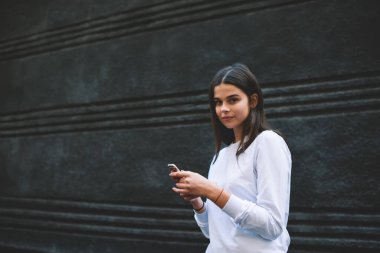 Uzun siyah saçlı, dost canlısı genç bir kadın. Sıradan bir kazak gibi giyinmiş. Modern cep telefonunda sörf yapıyor ve siyah duvara yaslanmış kameraya bakıyor.