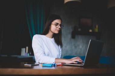Genç kadın gözlüklü ve günlük kıyafetleriyle modern çalışma alanındaki ahşap masada oturuyor ve ekrana bakarken dizüstü bilgisayarına bakıyor.