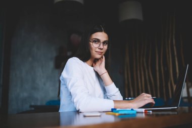 Genç, koyu renk saçlı, gözlüklü bir kız öğrenci netbook ile masada oturuyor ve kameraya gülümseyerek bakarken boynuna dokunuyor.