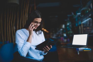 Gözlüklü ve dişlek gülüşlü pozitif kadın iş sırasında telefon görüşmesi yaparken ve not defterinden bilgi okurken aşağı bakıyor.
