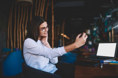 Yandan gülümseyen, serbest giyinmiş, gözlüklü, cep telefonlu, kafede dizüstü bilgisayarlı bir şekilde selfie çeken genç bir kadın görülüyor.