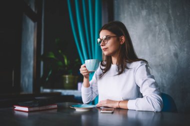 Genç düşünceli bir kadının kafeteryada dinlenip kahve içmesi ve kafeteryada akıllı telefon ve günlükle oturması.