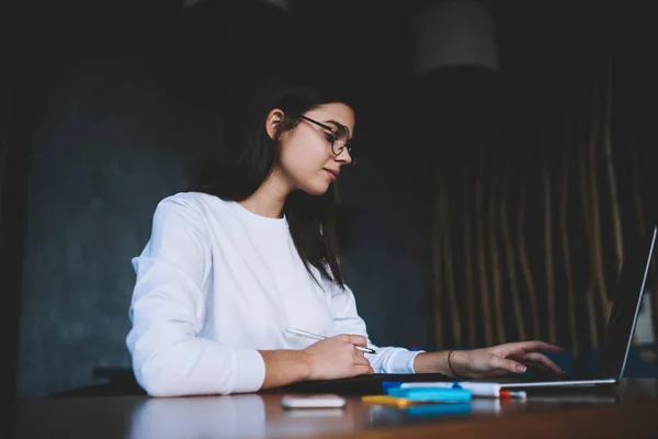 工作时 戴着眼镜 专心致志的女自由撰稿人与办公用品一起坐在木制桌子旁 浏览上网本的角度低 — 图库照片