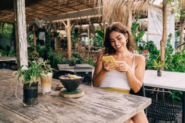 Genç içerikli etnik kadın ahşap masada cep telefonuyla sohbet ediyor verandada lezzetli yiyecekler ve kahveyle tropik tatil beldesinde.