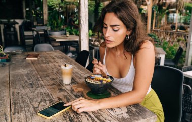 Tropikal tatil beldesindeki açık hava kafeteryasında bir kase lezzetli yemek ve kahveyle masada otururken konsantre kadın sörf cep telefonu.
