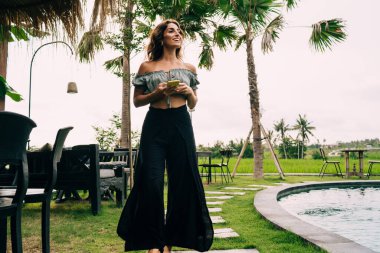 Moda kıyafetleri içinde gezen, cep telefonlarını karıştıran ve tropikal tatil beldesinde havuz kenarında yürürken gülümseyen genç, pozitif etnik bir bayan gezginin alçak açısı.