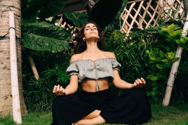 Barışçıl, genç, formalı, şık yaz kıyafetli, tropikal bahçedeki yeşil çimlerde oturan ve yarım Lotus yoga pozunda meditasyon yapan bir kadın.