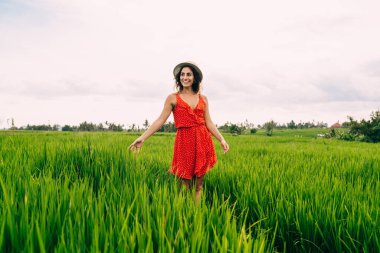 İyimser kadın, bulutlu bir günde çimenlikte yürüyen parlak kırmızı elbiseli ve gülümseyerek bakışlarını kaçıran.