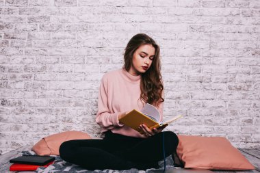 Uzun saçlı, rahat giyinen, bacak bacak üstüne atmış kitap okuyan, aydınlık odada vakit geçiren düşünceli genç bir kadın.