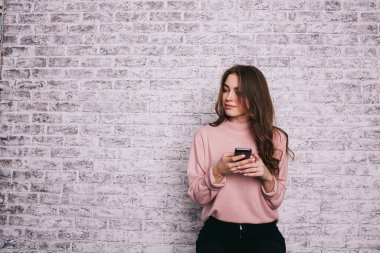Uzun saçlı, sıradan giysili genç bir bayan duvara bakıyor ve akıllı telefondan arkadaşlarıyla sohbet ediyor.