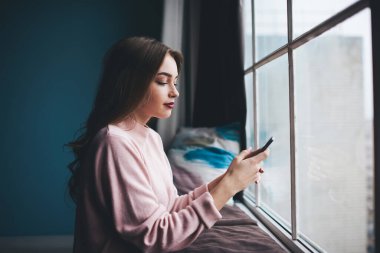 Uzun dalgalı saçları olan, akıllı telefondan arkadaşlarıyla sohbet ederken pencereye dayanan, günlük kıyafetli genç bir kadının yan görüntüsü.