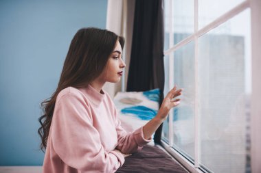 Pencereden dışarı bakan ve cama dokunan, uzun saçlı, sıradan giysili, düşünceli genç bir kadın.