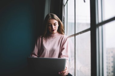 Ciddi kadın serbest çalışan, gündelik giysiler içinde, kırmızı dudaklı, pencerenin yanında dizüstü bilgisayarla oturmuş, uzaktan yeni bir proje üzerinde çalışıyor.