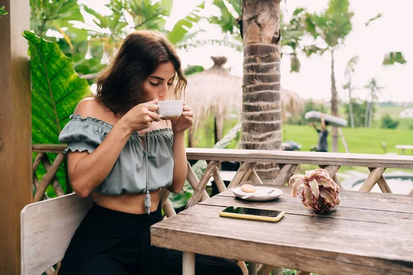 流行の衣装を着た若い民族の女性は 熱帯の国で休んでいる間 スマートフォンでテーブルに座ってカフェのテラスで熱いコーヒーを飲んでいます — ストック写真