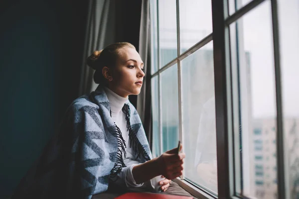 窓の外を見ながら窓にもたれかかる毛布に包まれた若い女性の側面図家で休息をしている — ストック写真