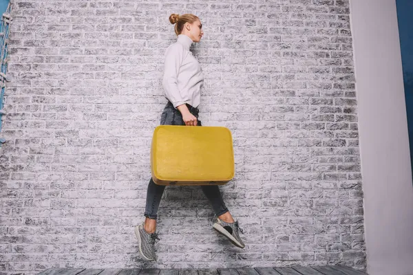スタジオでのレンガの壁の背景に黄色のスーツケースで飛び跳ねる離れて見てカジュアルな服装での高価な女性モデルの側面図 — ストック写真