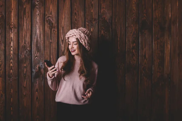 スマートフォンの画面を見て 木製の壁の近くの友人とチャットし おもちゃの笑顔でカジュアルな服装で若い幸せな女性 — ストック写真