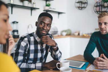 Genç erkek çalışan ve günlük kıyafetler içinde Afrikalı Amerikalı iş arkadaşı çalışma masasında otururken bayan meslektaşını dinliyor.