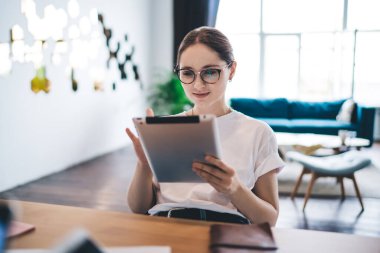 Gündelik giysiler ve gözlükler içinde mutlu genç bir bayan modern ışık odasında çalışırken masada oturuyor ve tablet okuyor.