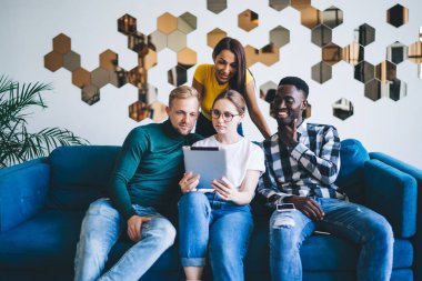 Çok ırklı iş arkadaşları gündelik giysiler içinde koltukta oturup tablet okuyorlar. Modern apartmanda birlikte çalışıyorlar ve duvarlarında yaratıcı süslemeler var.