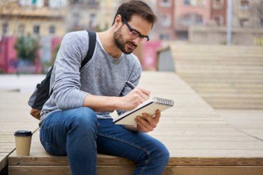 Yoğunlaşmış bir adam elinde bir fincan sıcak kahveyle dışarıdaki ahşap yapının üzerinde oturuyor ve iş sırasında yeni bir makale yazıyor.