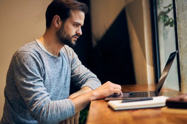 Rahat işyerindeki ahşap tezgahta oturan ve netbook üzerine yeni bir ödev üzerinde çalışan meşgul erkek yazarın yan görüntüsü