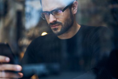 Modern kafede oturan ve cep telefonlarını karıştıran gündelik kıyafet ve gözlüklü ciddi bir erkeğin penceresinden.