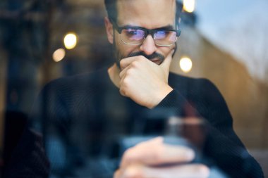 Ekin, etnik sakallı, sıradan giyinmiş, gözlüklü, modern kafede oturan ve akıllı telefondan sörf yapan düşünceli bir adamın penceresinden.