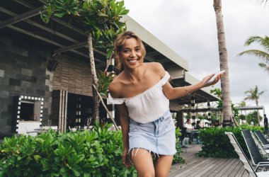 Yazlık bluzuyla neşeli genç bir bayan ve terasta yürüyen kot etekli akıllı telefonlu ve kameraya bakarken gülümseyen.