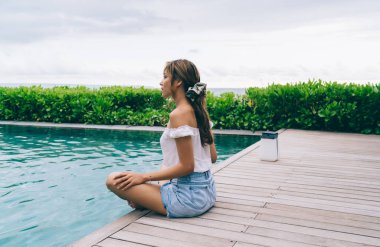 Nilüfer çiçeğinde oturan genç ve ince etnik kadının dış yüzme havuzunun yanında poz vermesi ve yaz tatili boyunca tropikal tatil boyunca meditasyon yapması.
