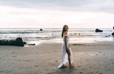 Beyaz plajlı genç, etnik bir bayanın tropik tatil köyünde gün batımında boş okyanus sahillerinde tek başına dolaşmasının yan görüntüsü.