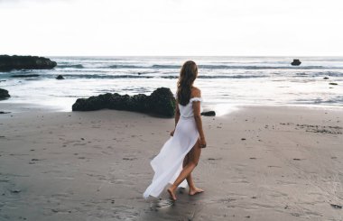 Yüzü olmayan, bronz tenli, beyaz elbiseli bir gezginin yaz tatilinde ıslak kumda tek başına yürümesi.