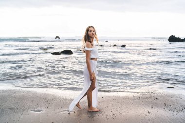 Kahverengi saçlı, beyaz omuzlu, açık etekli, kumlu sahilde su kenarında yürüyen düşünceli genç bir kadın.