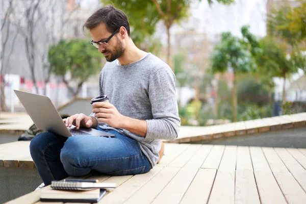 Side view of focused male freelancer searching information on computer while spending time in street and drinking hot beverage during work