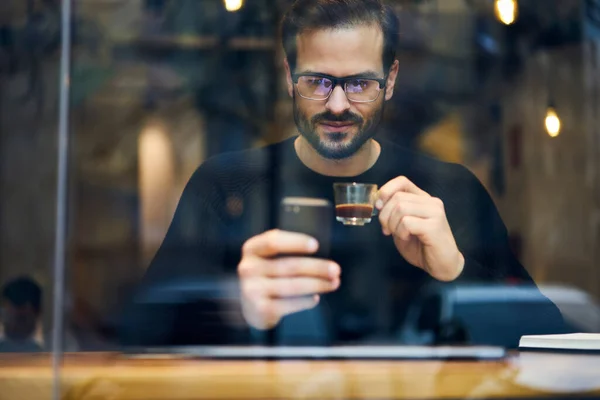 Through window of bearded ethnic male in casual wear and glasses with cup of hot coffee sitting in cafe and surfing smartphone