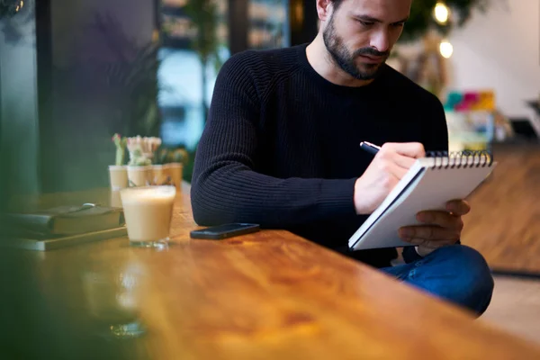 作物集中长胡子的男性 穿着休闲装 坐在木制桌旁 一边远程工作一边在抄本上记笔记 — 图库照片
