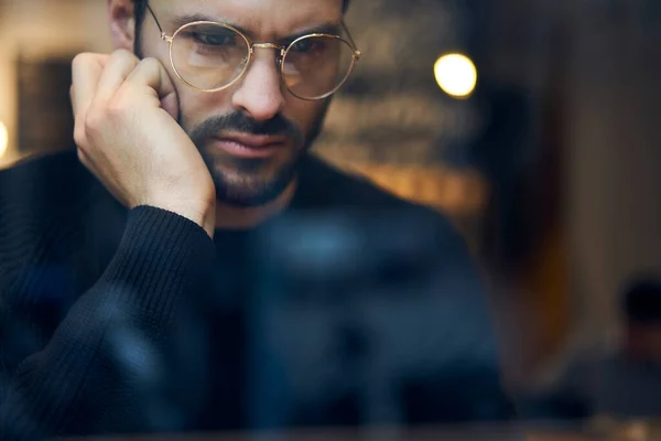 Though window low angle of crop bearded ethnic serious male manager in casual outfit and glasses working on laptop remotely