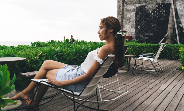 Yazlık giysiler içinde çıplak ayaklı bir kadın otelin verandasındaki koltukta dinleniyor ve tatilin tadını çıkarırken denize bakıyor.