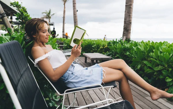 縁側の上のラウンジャーでリラックスし 熱帯リゾートで夏休みの間に興味深い本を読んで肯定的な若い民族の女性の側面図 — ストック写真