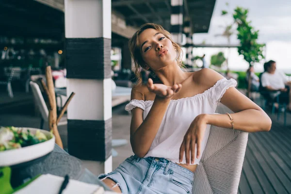 Modern bir açık hava restoranında açık omuzlu, beyaz bluzlu, pozitifti. Kameraya bakarken hava öpücüğü gönderiyordu.