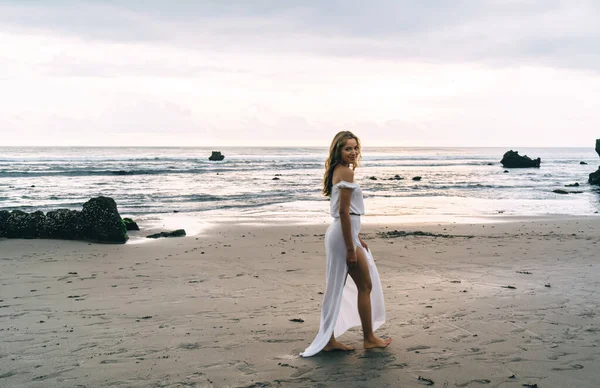 Beyaz plajlı genç, etnik bir bayanın tropik tatil köyünde gün batımında boş okyanus sahillerinde tek başına dolaşmasının yan görüntüsü.