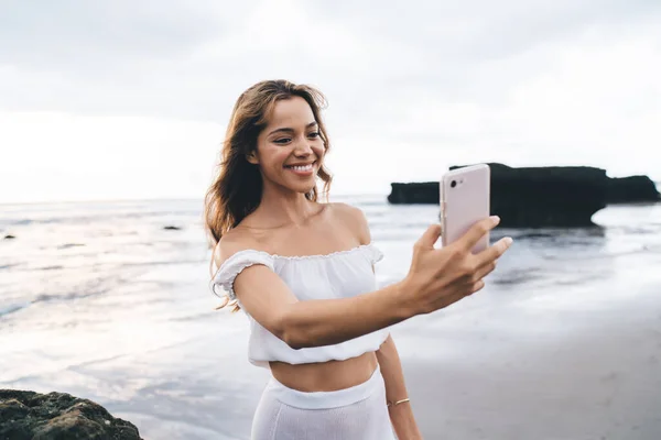 年轻快乐的女性穿着白色的夏装站在沙滩上 用手机拍着大海和岩石 — 图库照片
